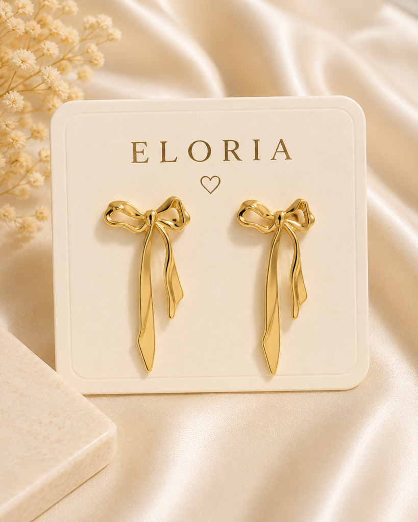 Gold-tone bow ribbon drop earrings displayed on an ELORIA jewelry card over a luxurious satin background.
