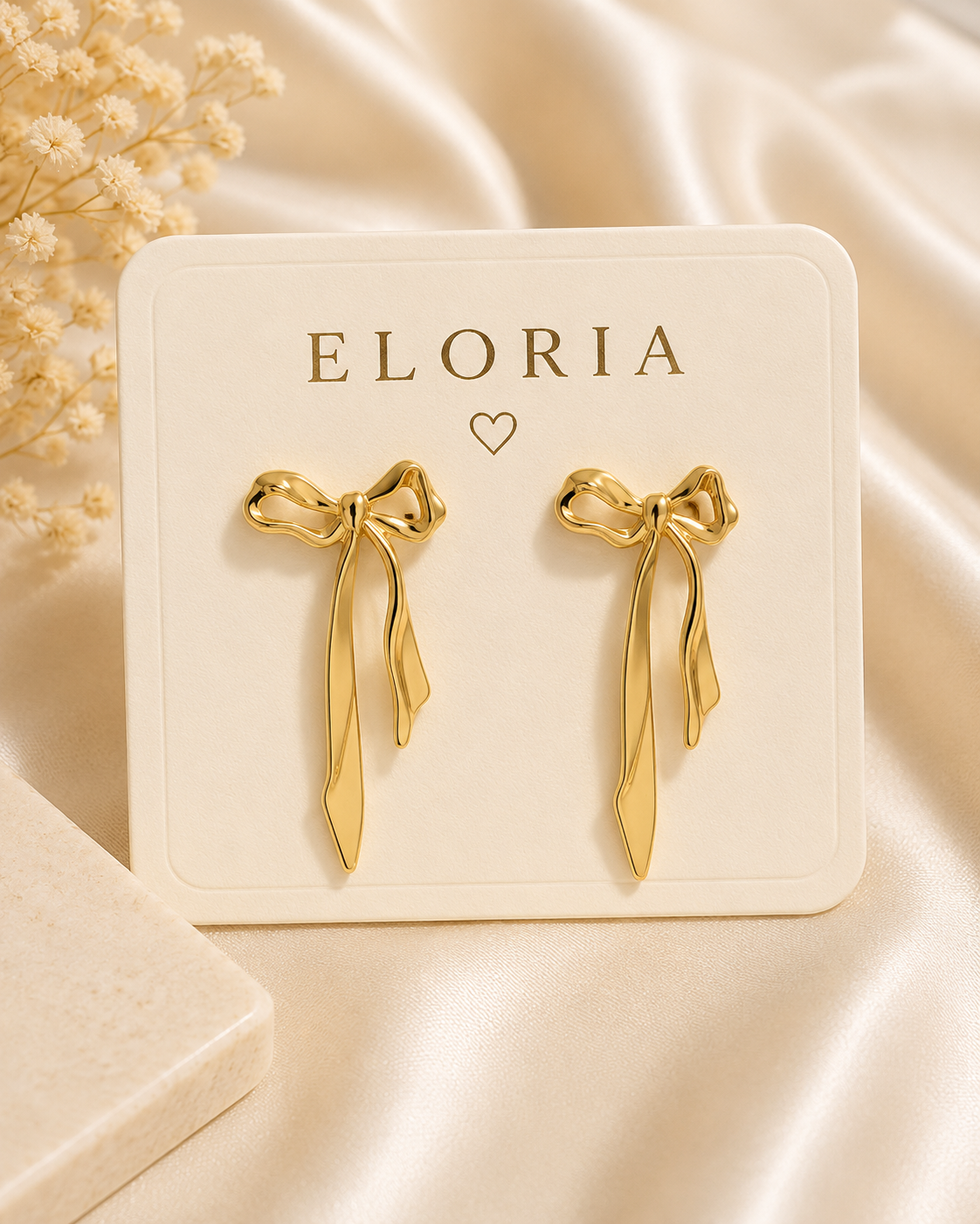Gold-tone bow ribbon drop earrings displayed on an ELORIA jewelry card over a luxurious satin background.