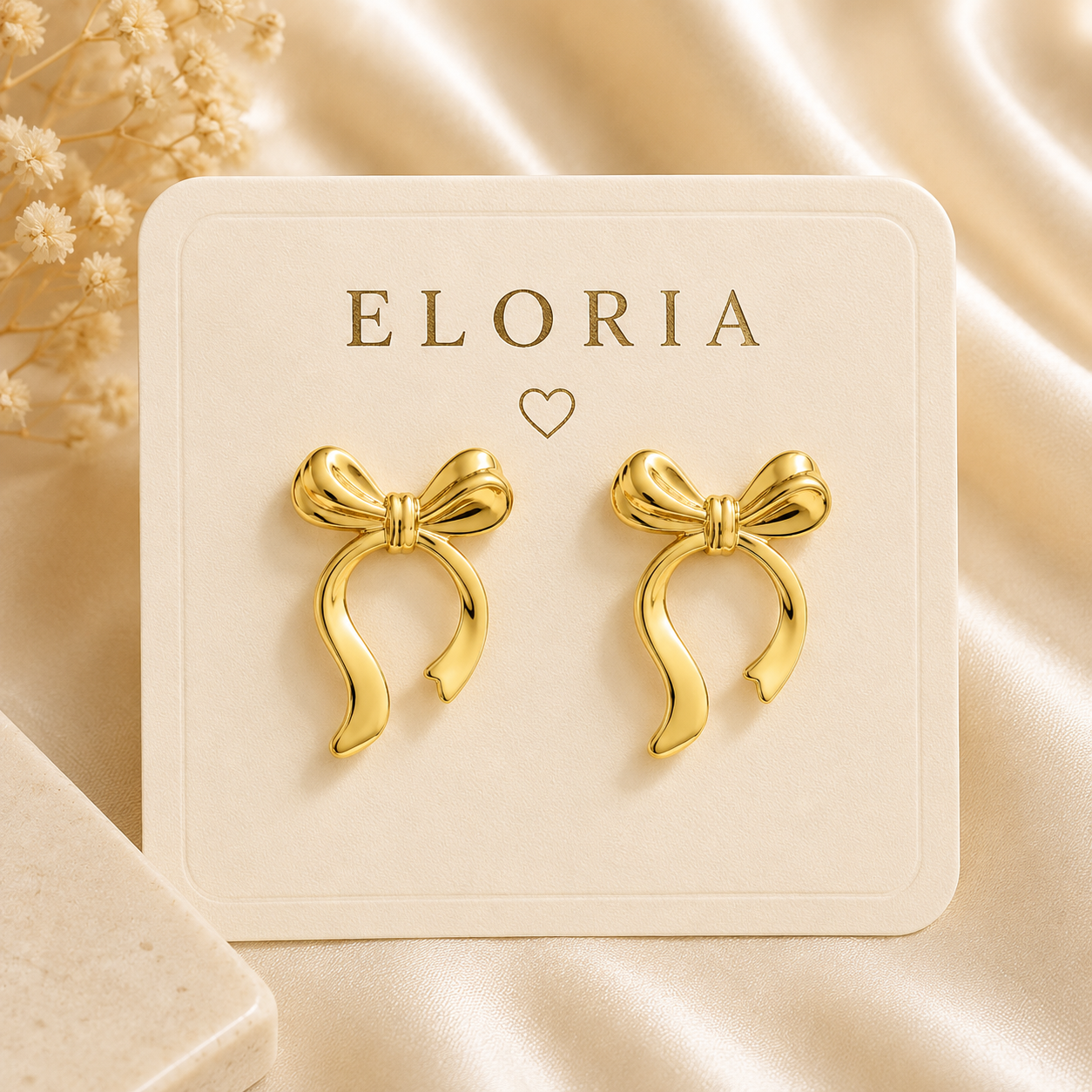 Gold-tone bow ribbon loop earrings displayed on a white jewelry card over champagne satin.